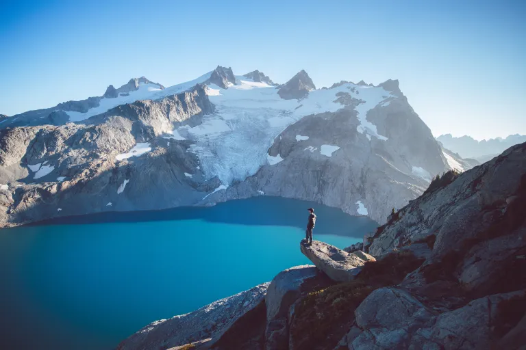 A turquoise blue lake surrounded by a mountain peak with snowfields. The foreground shows a man standing on a ledge overlooking the lake.