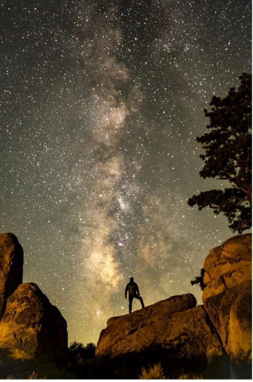 The Milky Way rises up the center of the photo from golden rocks and a person is silhouetted against the fiery night sky. 