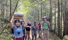 A diverse group of hikers on the trail, facing the camera with one hiker in front holder a bundle of campfire wood above them triumphantly