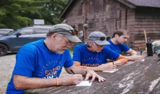 Three people write letters on a park bench outdoors. 