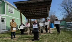 Five people stood outdoors in a small neighborhood park. One person is standing forward of the others and is speaking through a microphone. There is a sign on the microphone stand saying NO data center. The other people in the photo are all holding signs too. There area  few trees in the background which have shed their leaves. The sky is blue.