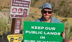 amn in sunglasses and hat crouched next to a trailhead sign holding a green sign with white text that says keep our public lands in public hands