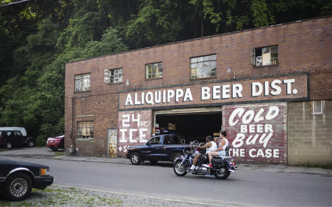 A man and woman on a motorcycle pass in front of a brick building with painted signs saying Aliquippa Beer Dist., 24hr Ice, and Cold Beer Buy the Case.