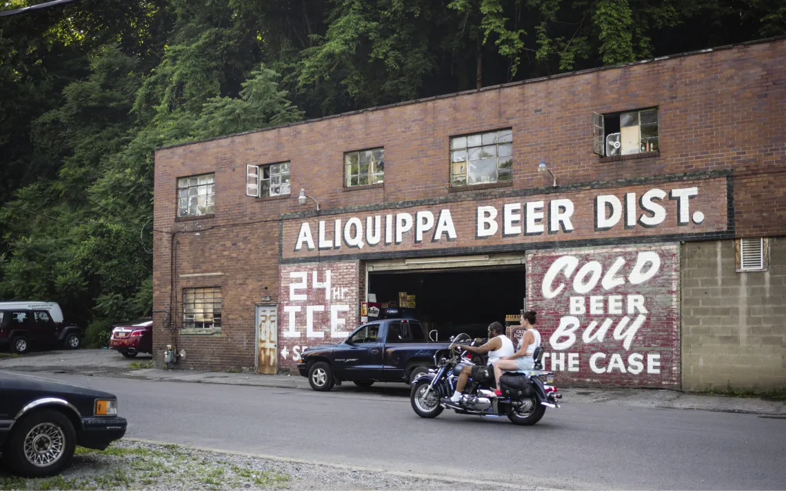 A man and woman on a motorcycle pass in front of a brick building with painted signs saying Aliquippa Beer Dist., 24hr Ice, and Cold Beer Buy the Case.