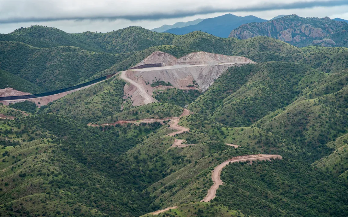 Aerial photo shows a section of border wall going through a green, hilly section. The side of a hill has been sliced off to fit the wall and several dirt roads cut through the greenery.