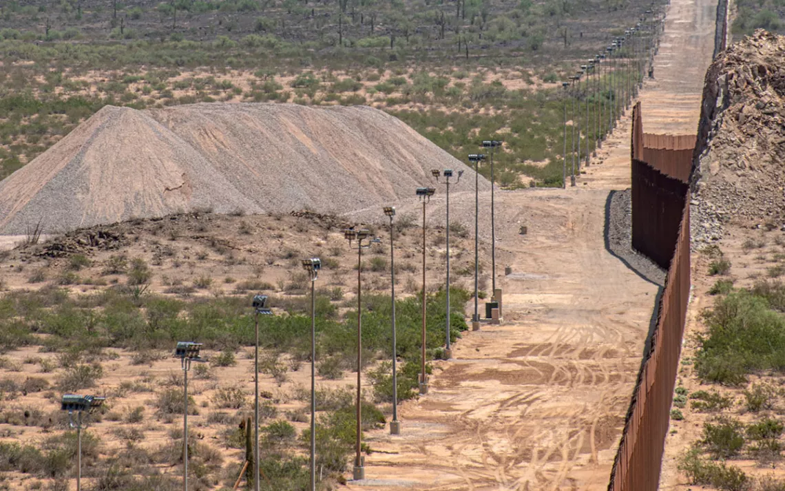 Close-up of the rust-colored border wall and a dirt road along with some cameras atop tall poles. There are some cacti and a pile of rock in the background.