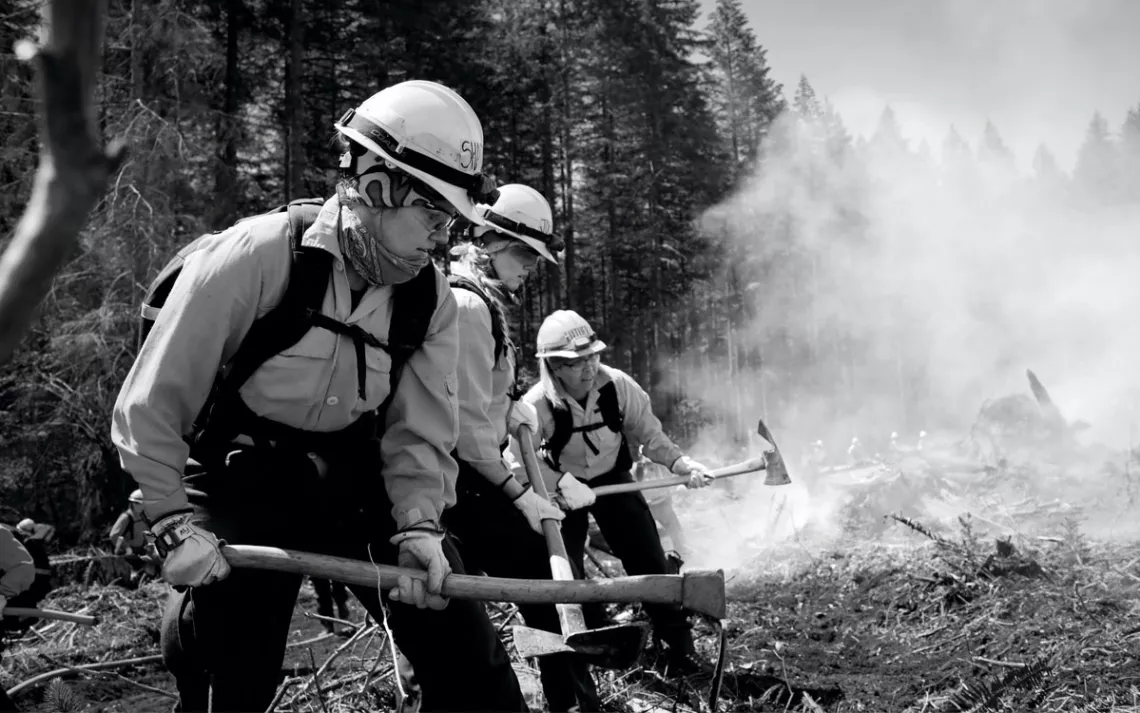 A row of female firefighters are suited up and holding axes to cut a fire line.