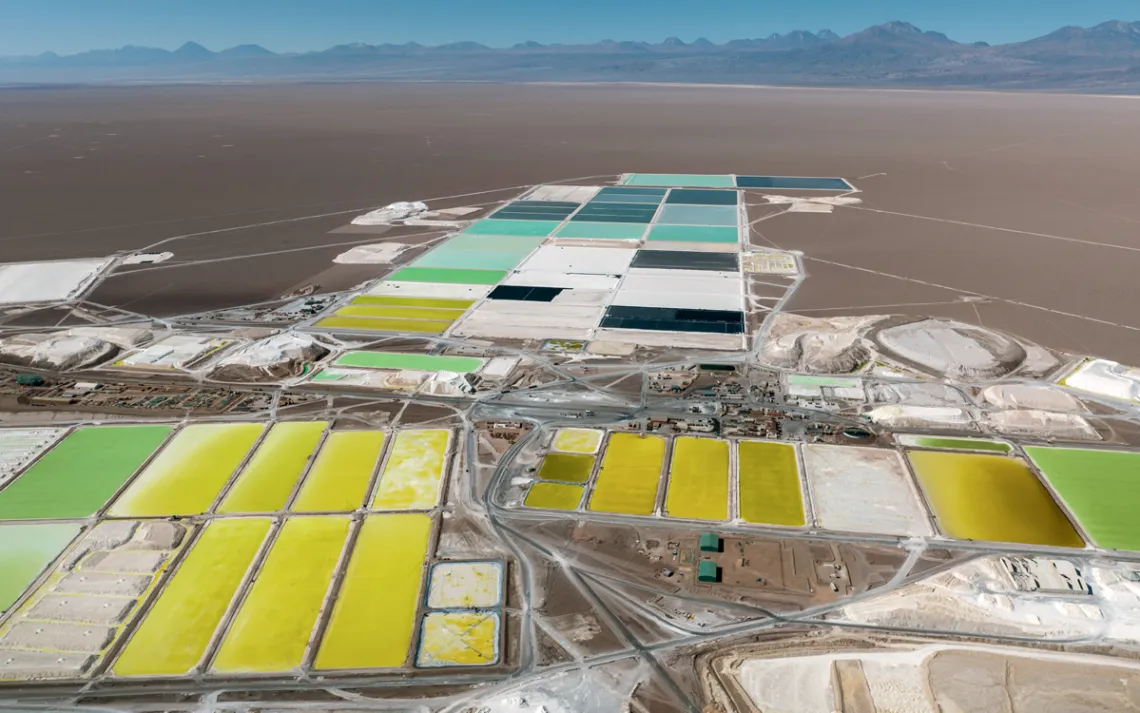 Aerial photo of a dark-sand desert with several shallow lithium pools in greens and blues and yellows.