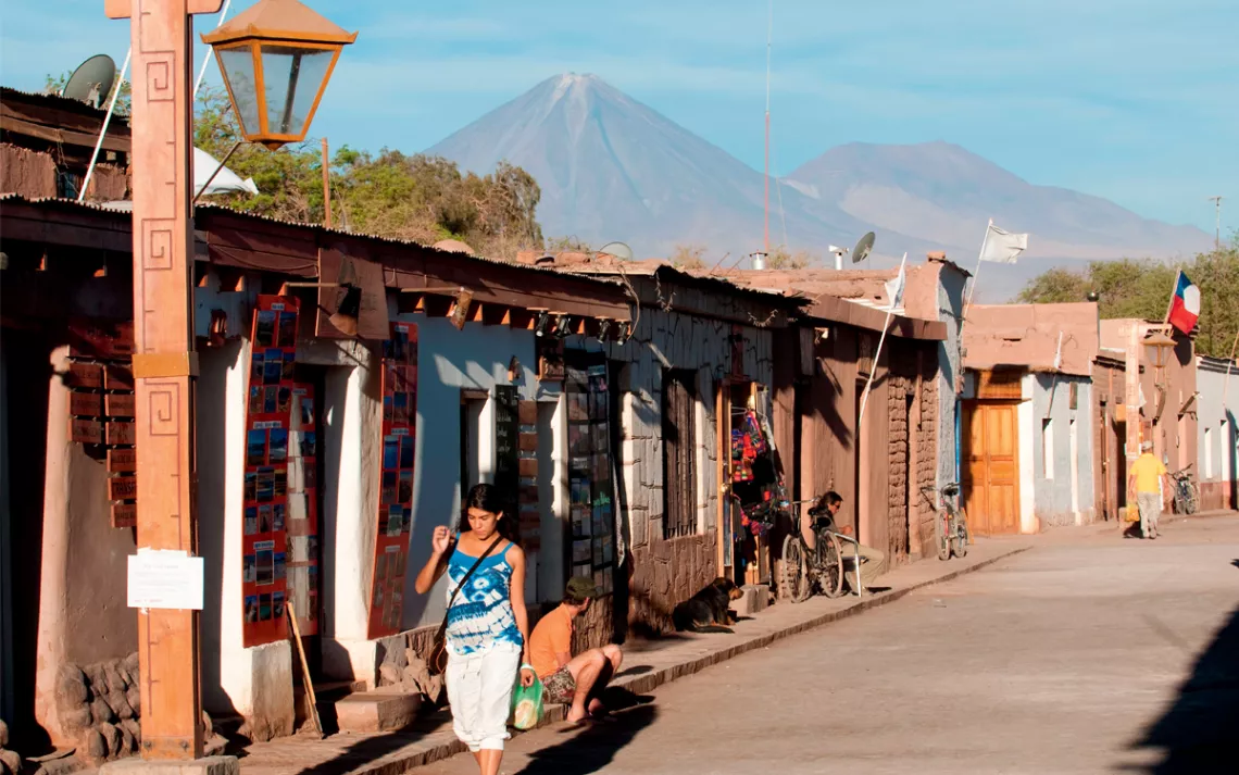 Two people are walking and two sitting next to a paved road and storefronts with mountains in the background.