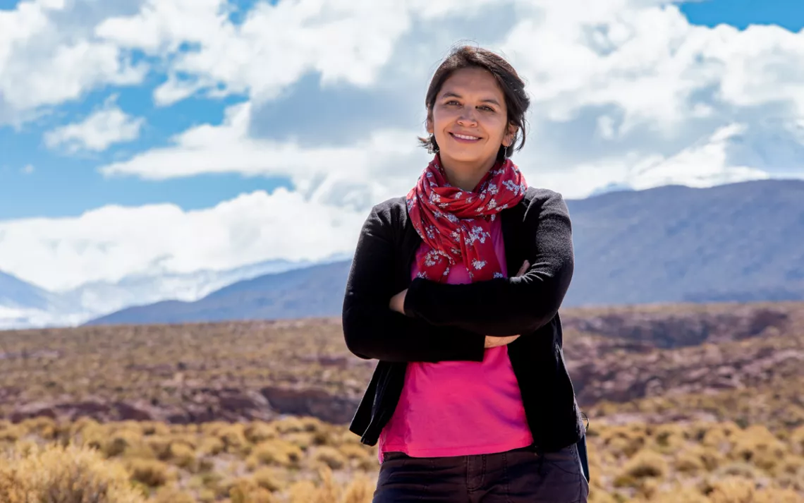 Cristina Dorador wears a pink shirt and scarf. She's smiling with her arms folded in front of her. Behind her is a mountain and a cloudy sky.