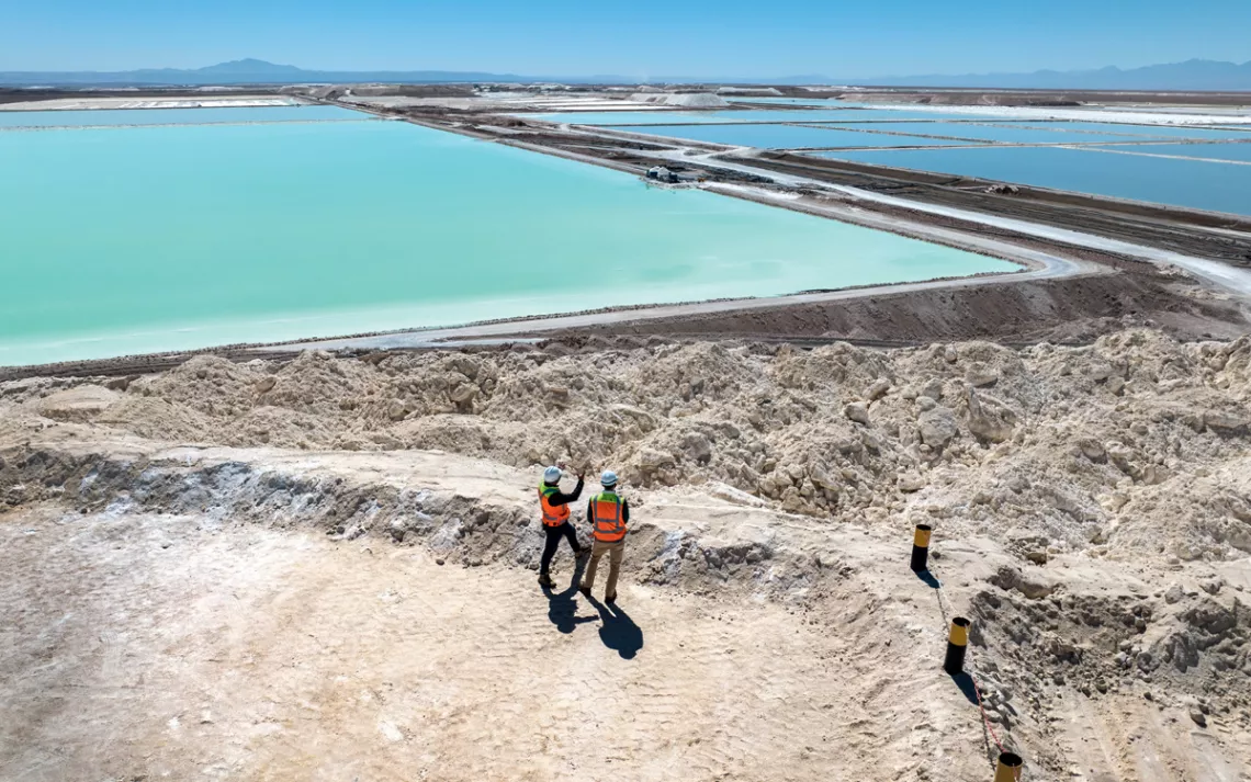 Two people wearing orange vests and hardhats look out at large blue lithium ponds.