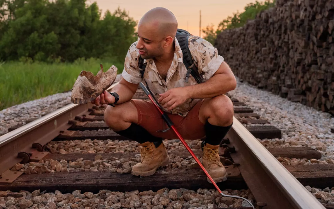 Alexi Grousis squats down in the middle of some train tracks. He has a metal hook tool and is holding up what looks like an old turtle shell.