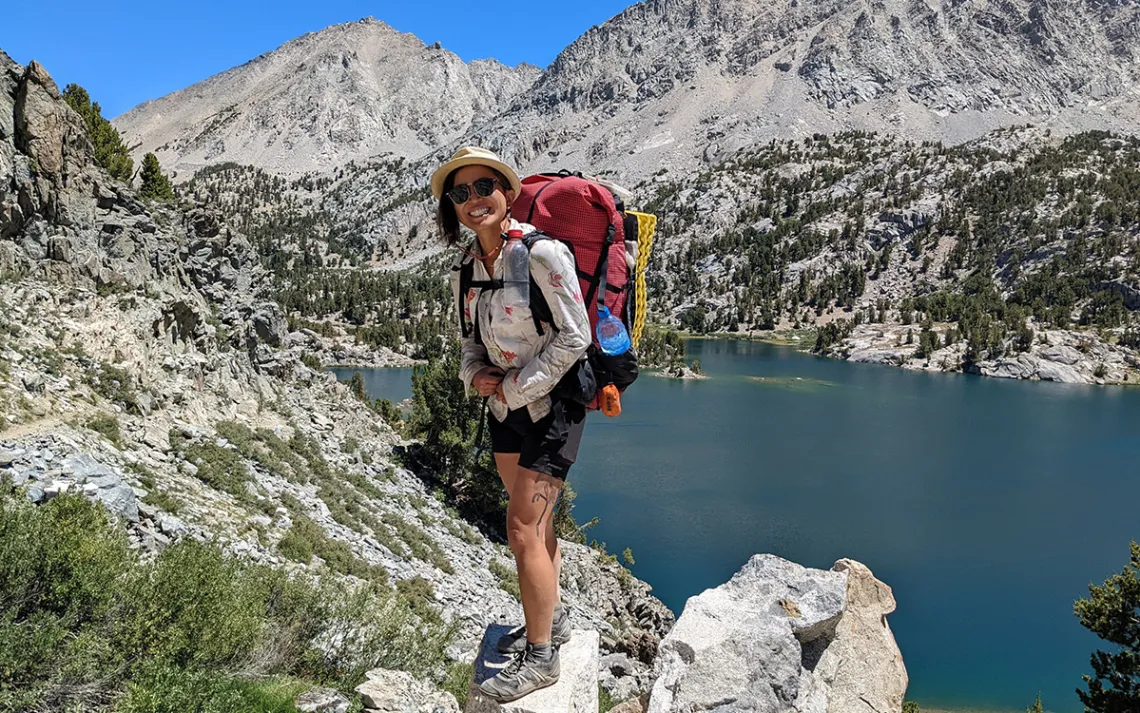 Image of Narinda Semiseki wearing a backpack and outdoor gear smiling in front of a mountainous landscape. 