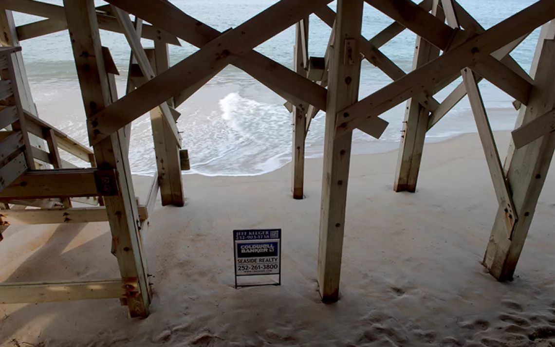 View looking through the stilts of a house toward the Atlantic Ocean, with waves lapping up to a Coldwell Banker real estate sign in the sand at Nags Head, North Carolina.