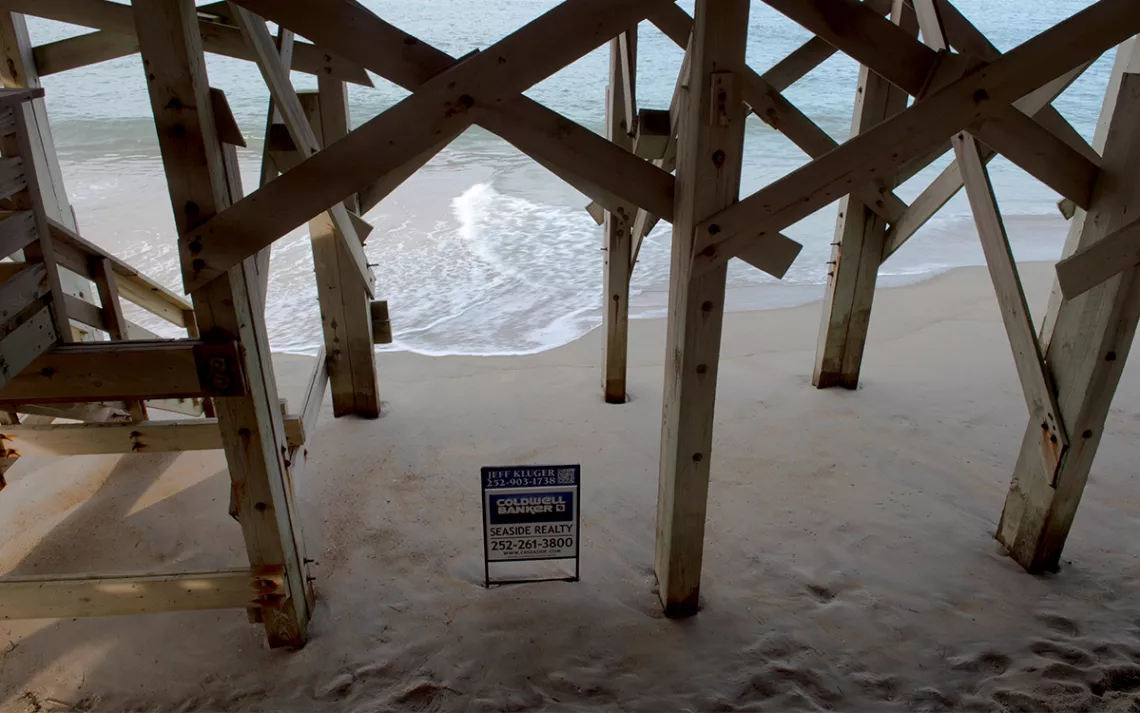 View looking through the stilts of a house toward the Atlantic Ocean, with waves lapping up to a Coldwell Banker real estate sign in the sand at Nags Head, North Carolina.