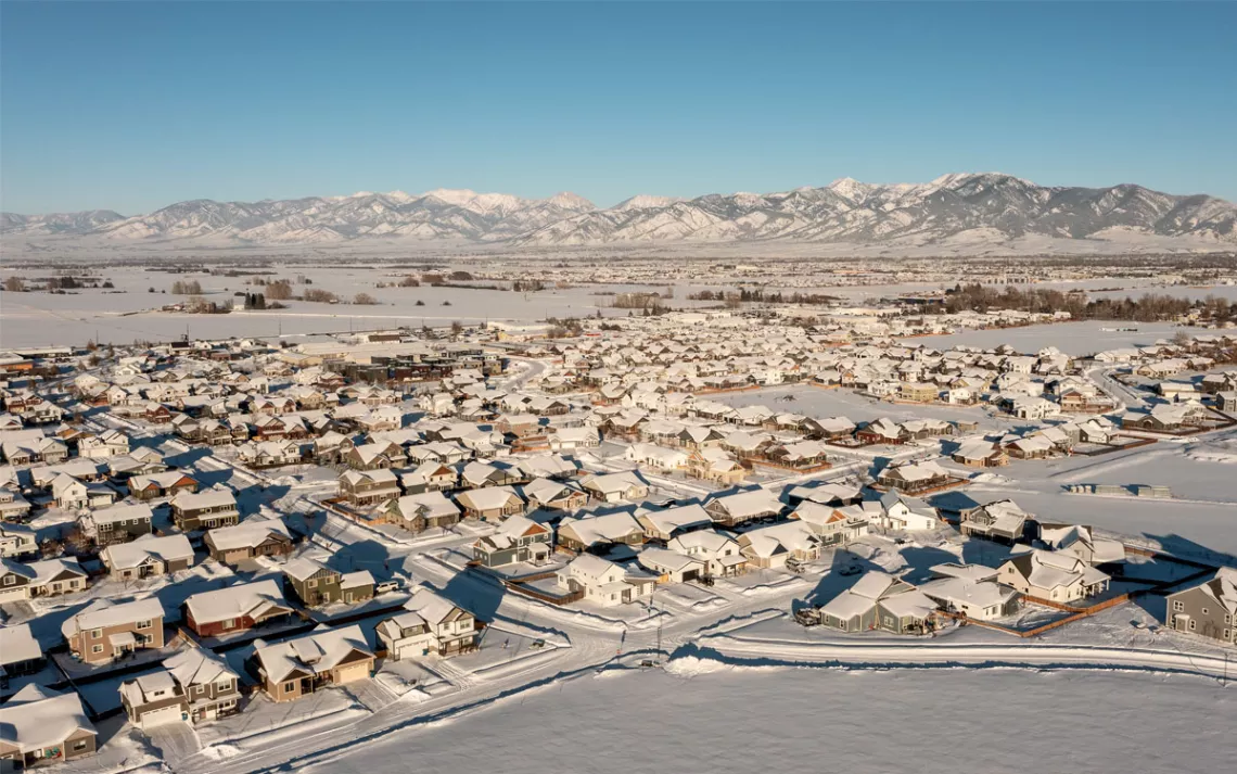 Aerial photo shows a flat housing subdivision covered in snow with mountains in the background.