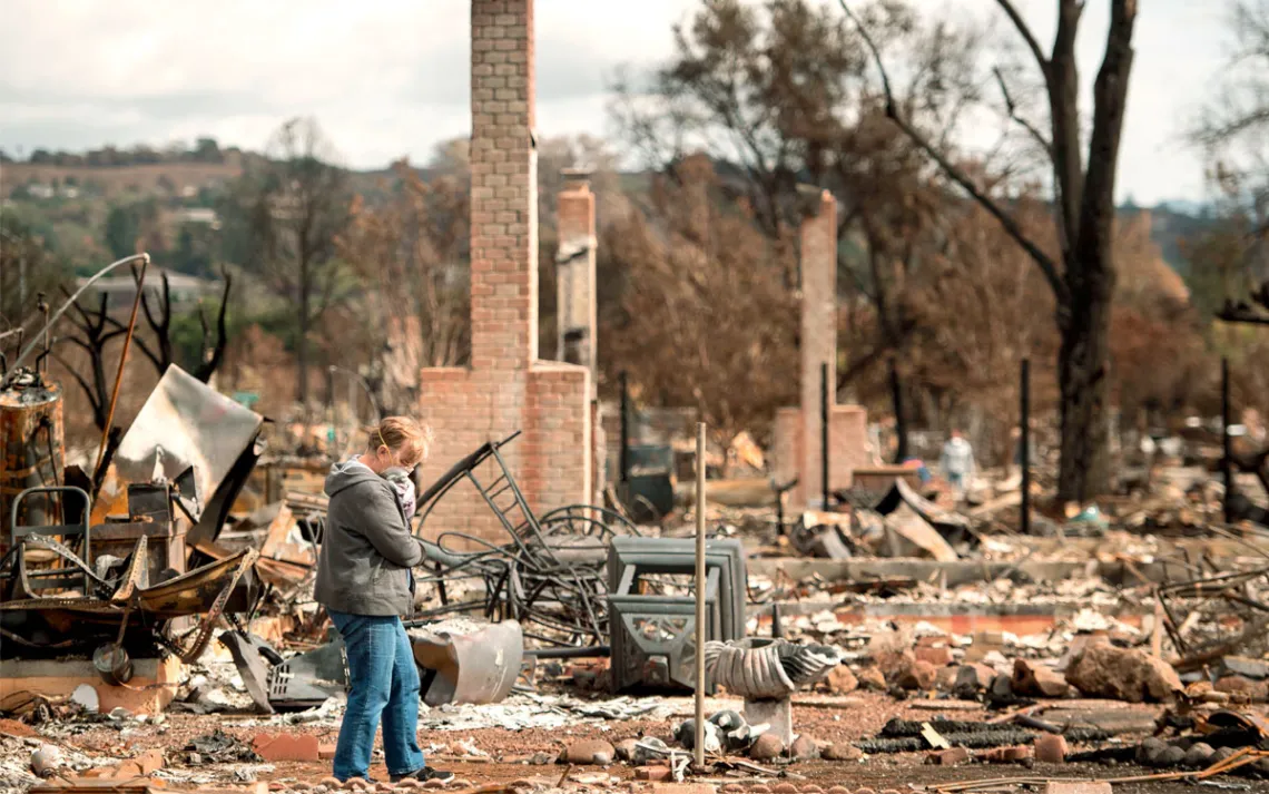 A woman stands next to the burned remains of a home, including a brick fireplace and metal items.