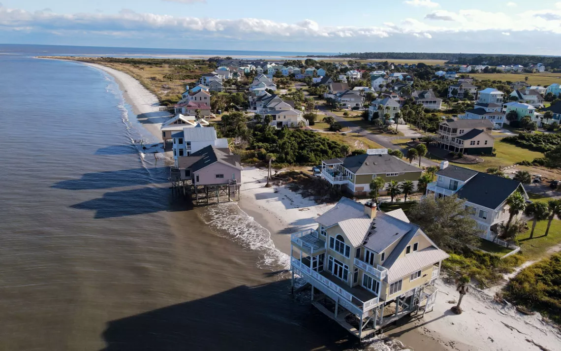 Aerial photo shows a beach with several houses on stilts sitting on the waterline.
