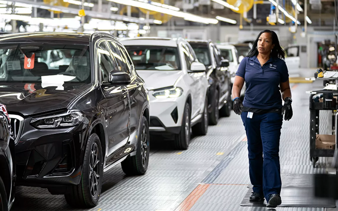 A female worker in all blue walks by a line of new cars in a factory.