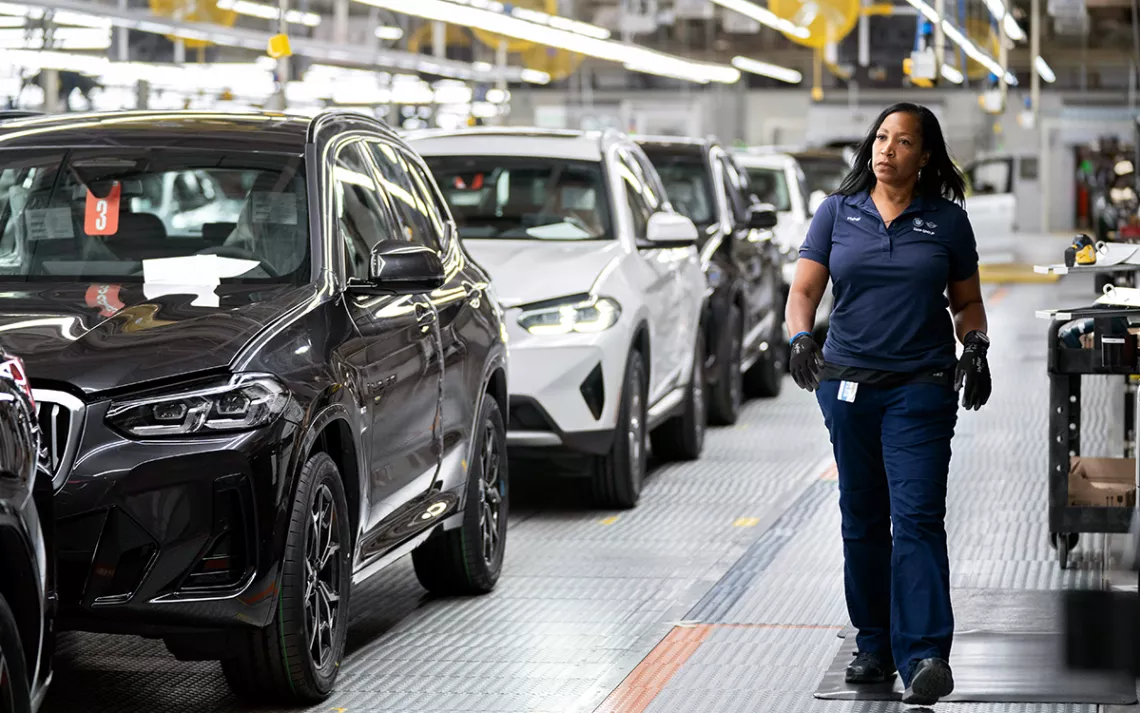 A female worker in all blue walks by a line of new cars in a factory.