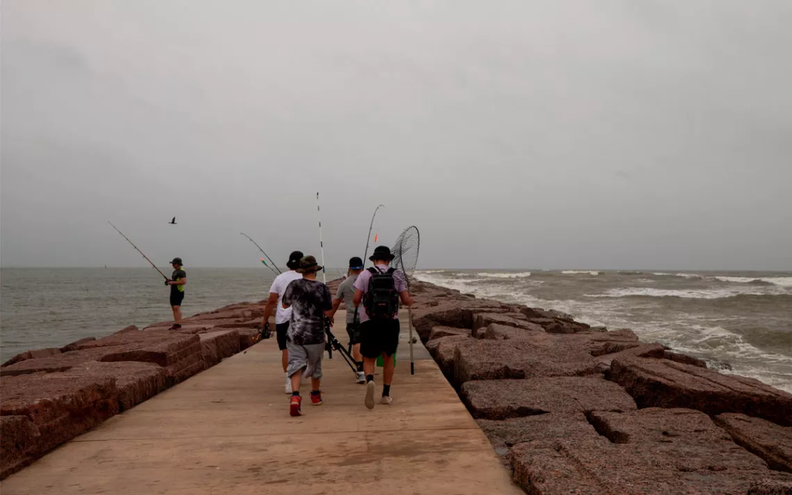 Five fishers are on a jetty with fishing poles on an overcast day.
