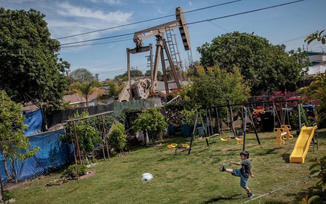 A kid is playing soccer in the backyard. Right behind the yard fence is an oil drill.