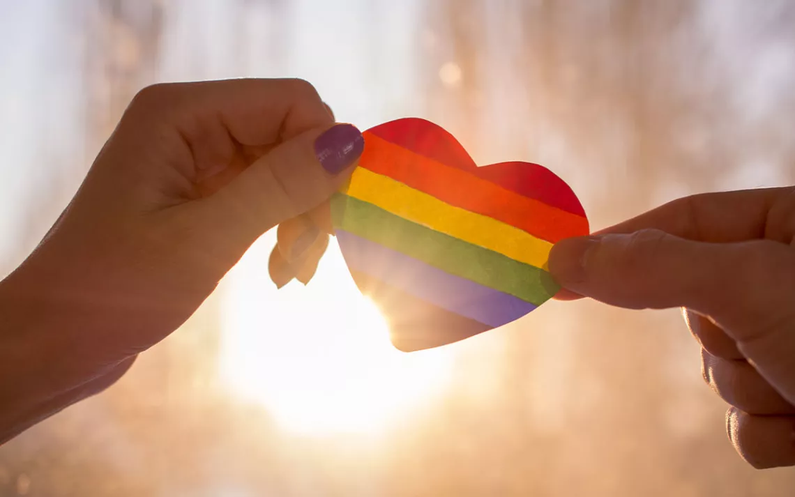 Hand hold a heart painted like a LGBT flag, silhouetted against sun