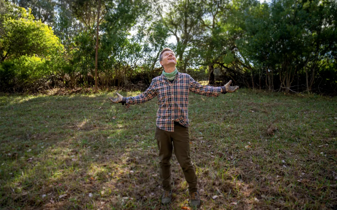 Ole Schell stands in a grassy field with a plaid shirt and his arms spread, smiling and looking at the sky.