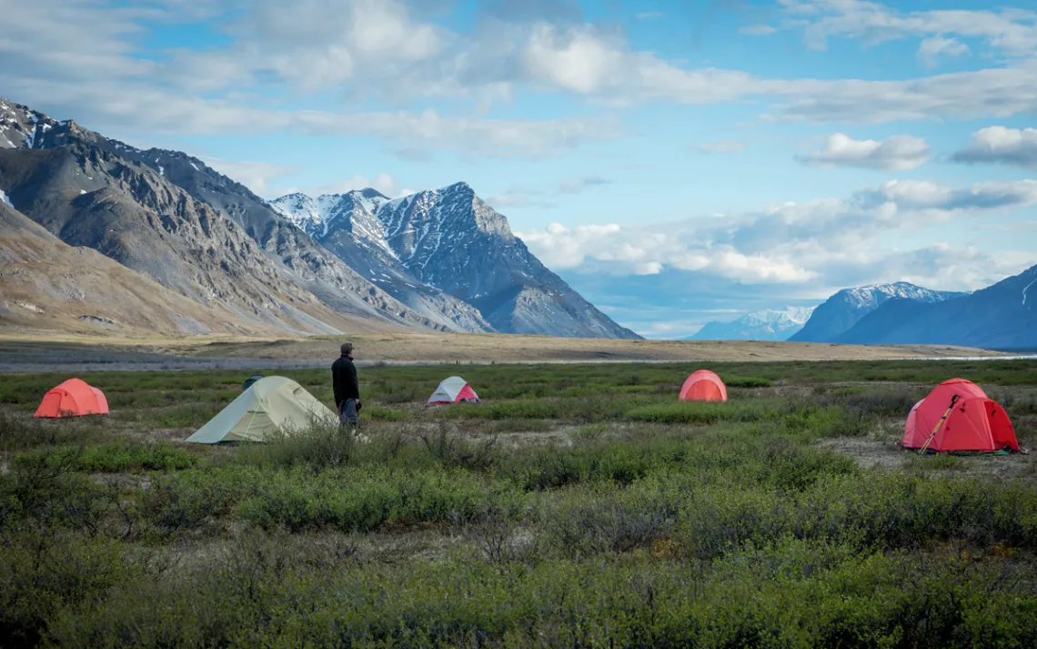  A man stands near four tents in an Arctic Refuge valley meadow with snowcapped mountains in the background.