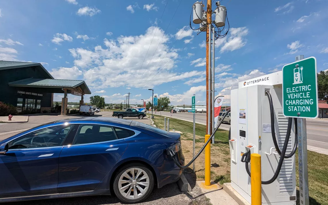A royal-blue Tesla is charging at an Otterspace station with a sign that reads "Electric Vehicle Charging Station" next to a store and a road.