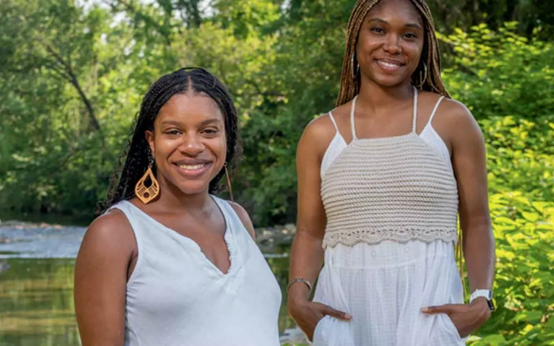 Diamon Clark and Kristen Walker stand by a tree-lined creek wearing long white dresses.