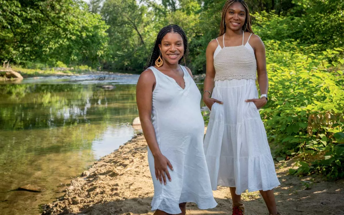 Diamon Clark and Kristen Walker stand by a tree-lined creek wearing long white dresses.