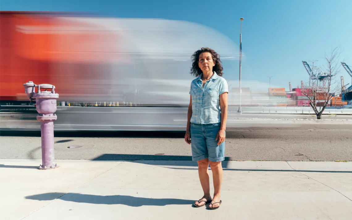 Alicia Rivera is wearing a light blue shirt, jean shorts, and flip flops and standing in front of traffic by the port.