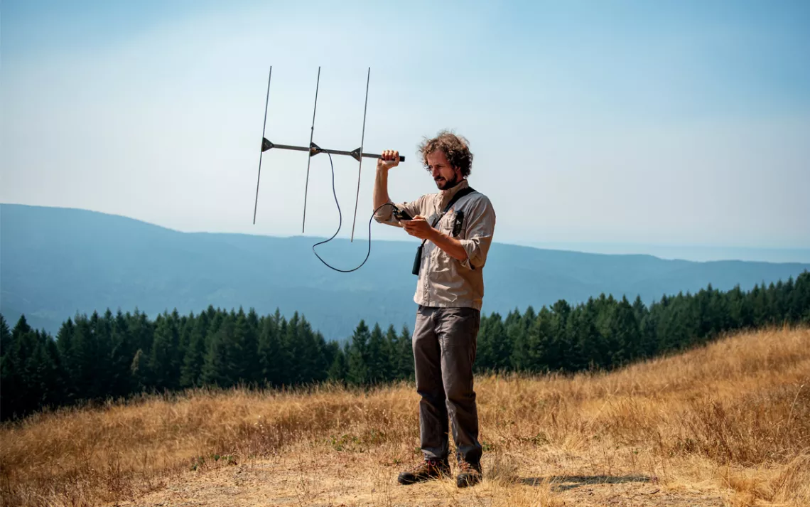 Patrick Myers stands in a dry grassy field atop a mountain and holds a telemetry device out in front of him.