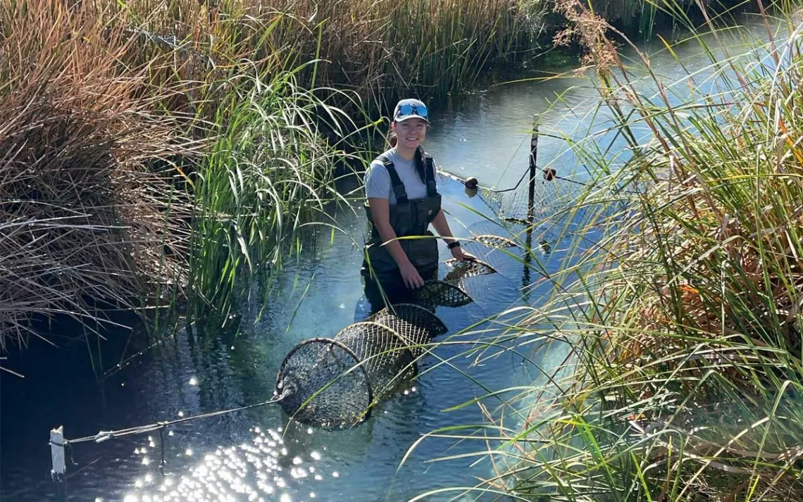  Emma Priger is wearing waders and smiling at the camera while holding a hoop net in a narrow tallgrass-lined stream.