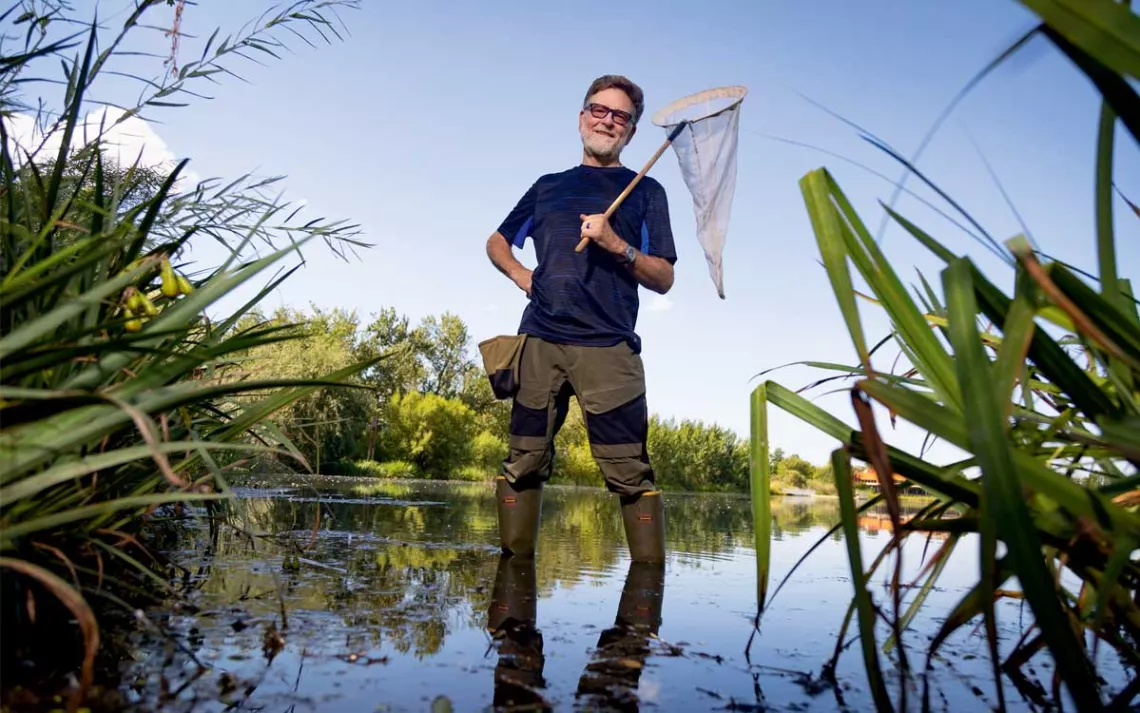 Dick Jordan smiles at the camera and holds a net while wearing tall wading boots and standing in a grass-lined body of water.
