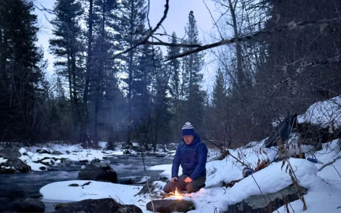 Aaron Teasdale, wearing a blue puffer jacket and ski cap, kneels down in snow in front of a small fire by a wooded creek.