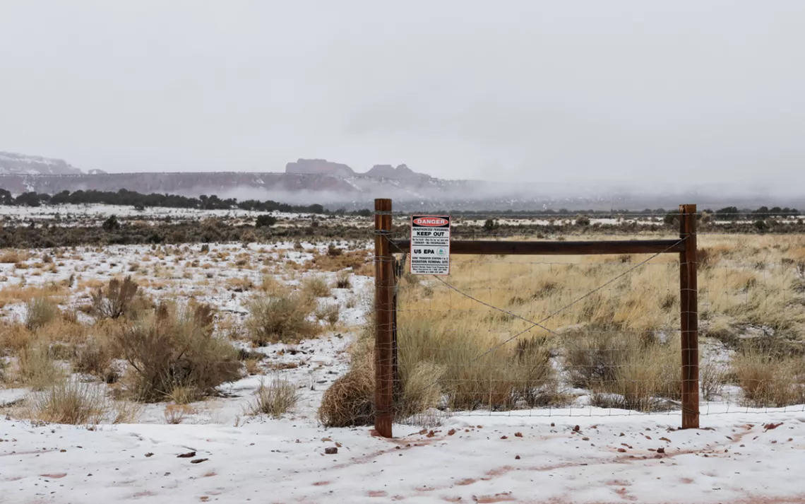 A barbed-wire fence in front of flat, shrubby terrain. A sign reads "Danger. Keep Out. US EPA Cove Transfer Station 1 & 2 Radiation Removal Site. Contact Navajo Super Fund program for more information."