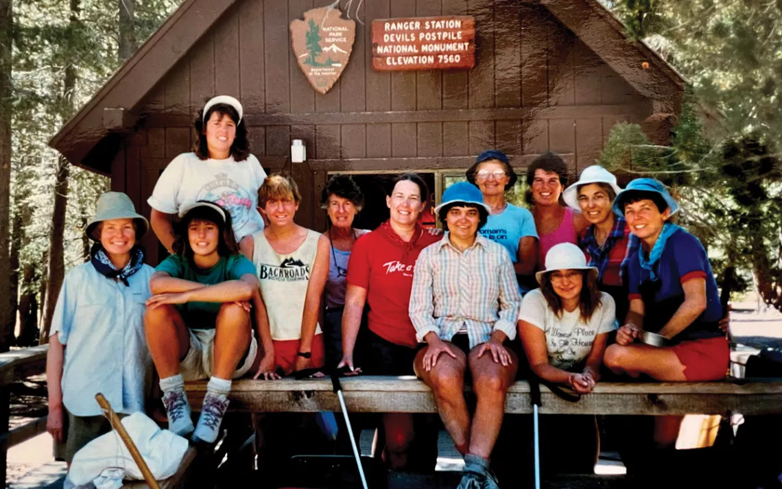 Twelve people stand in front of a brown wooden building in a wooded area with a sign that reads Ranger Station / Devils Postpile National Monument / Elevation 7560.