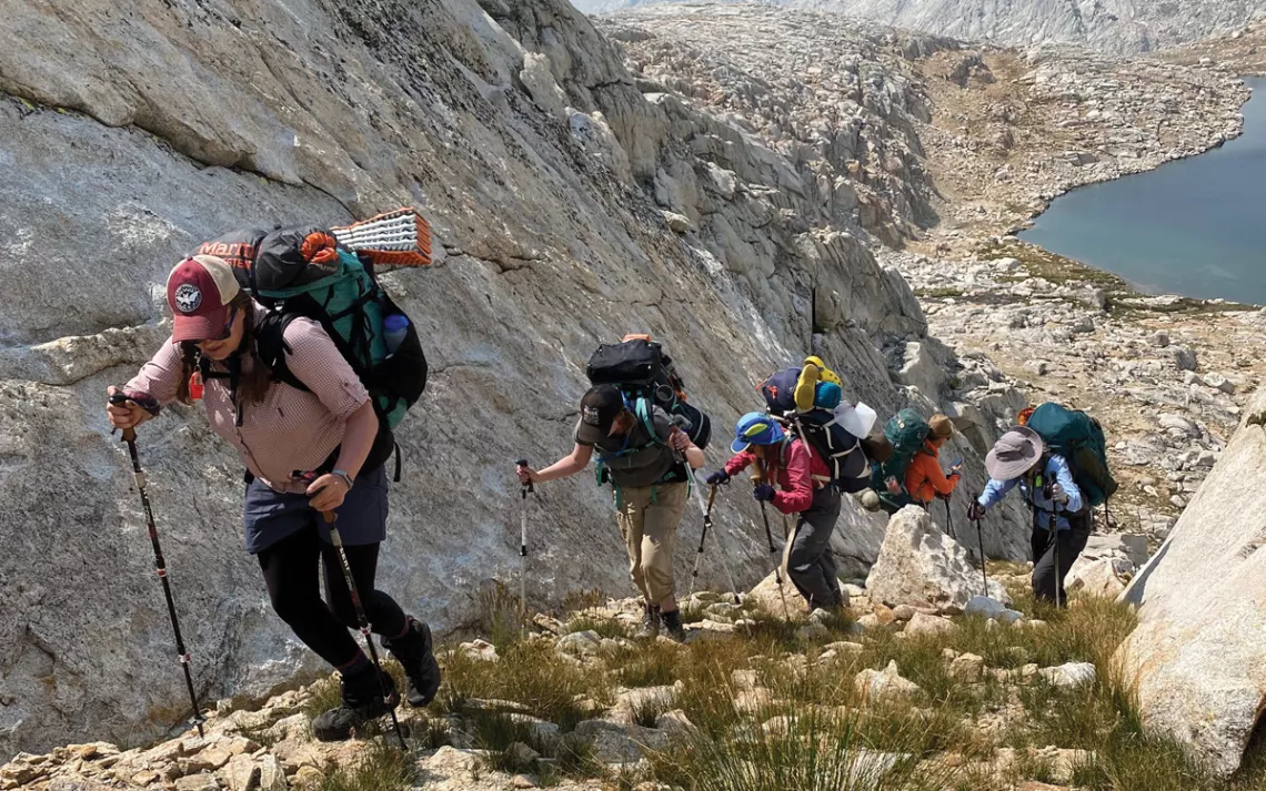 Five people climb a rocky mountainside with hiking sticks, with a lake below, in John Muir Wilderness.