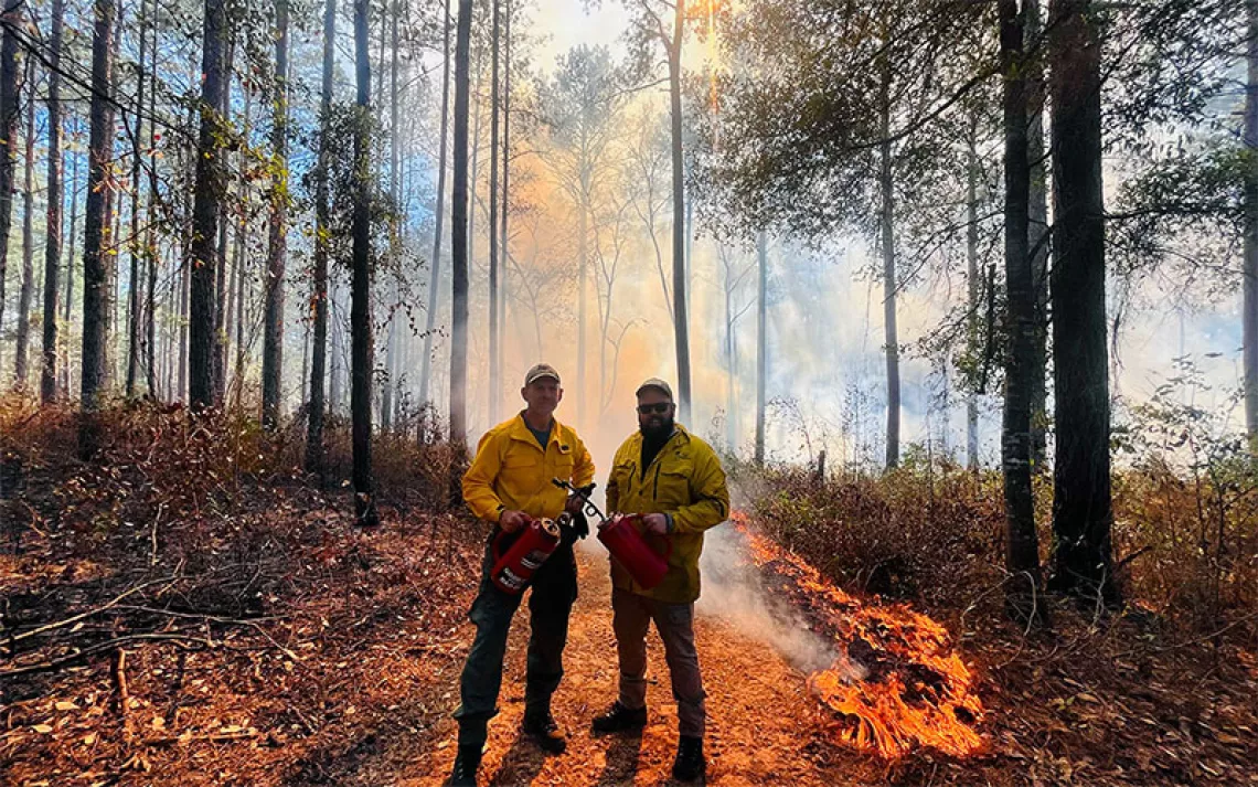 Two men dressed in yellow PPE and holding fuel canisters set a prescribed burn to a forest in Southwest Georgia.
