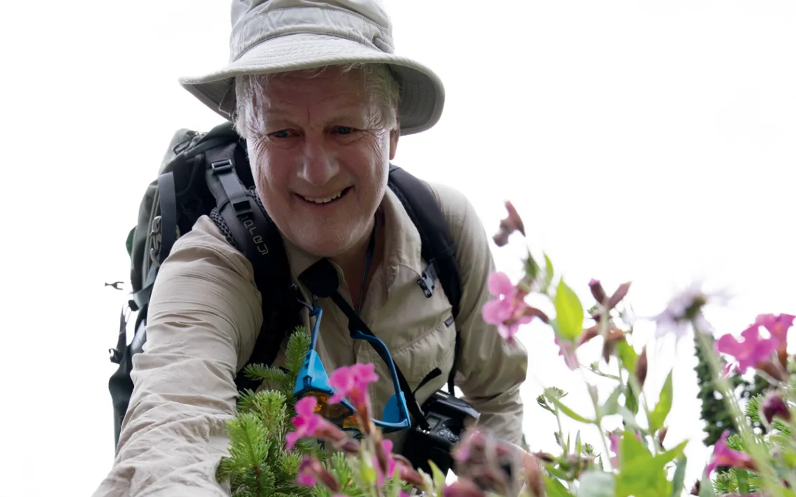 Harvey Locke, wearing a backpack and beige field hat, smiles and reaches his arm out toward some pink flowers.