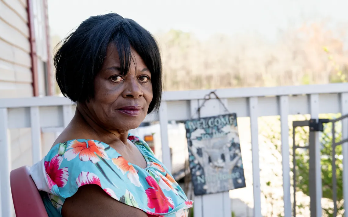 Elaine Howard wears a floral shirt and sits on a porch looking sideways at the camera