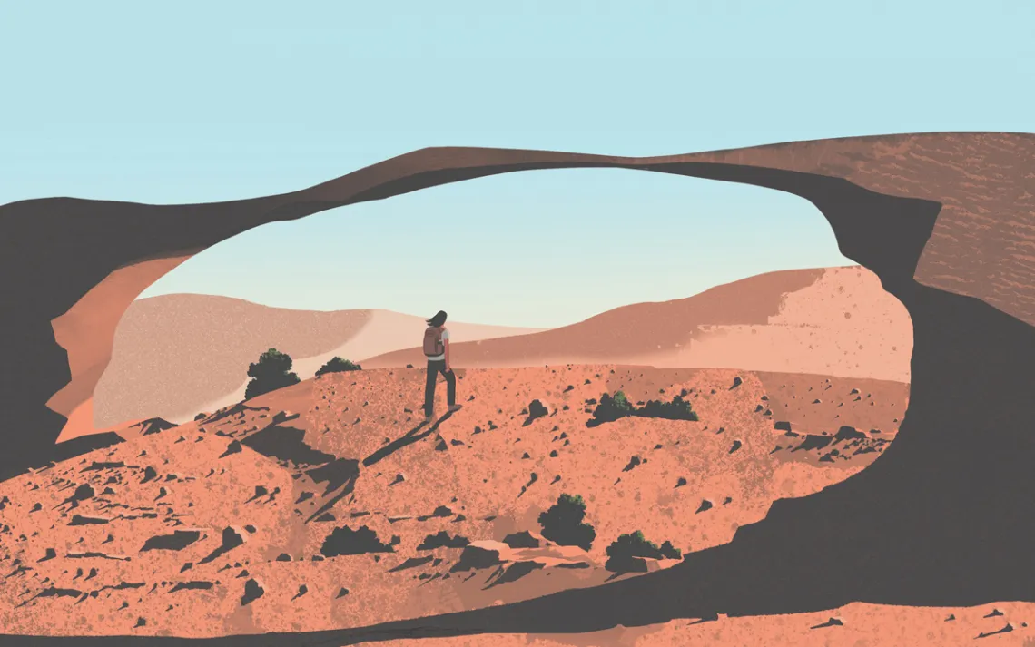  A hiker walks up a redrock hill toward a sandstone arch in the desert