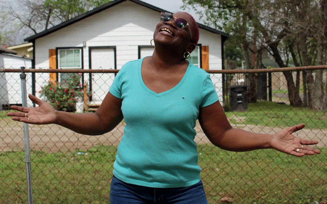 Sandra Edwards wears a turquoise shirt and sunglasses and holds her hands up to the sky and smiles. Behind her is a chain-link fence and a small white house.