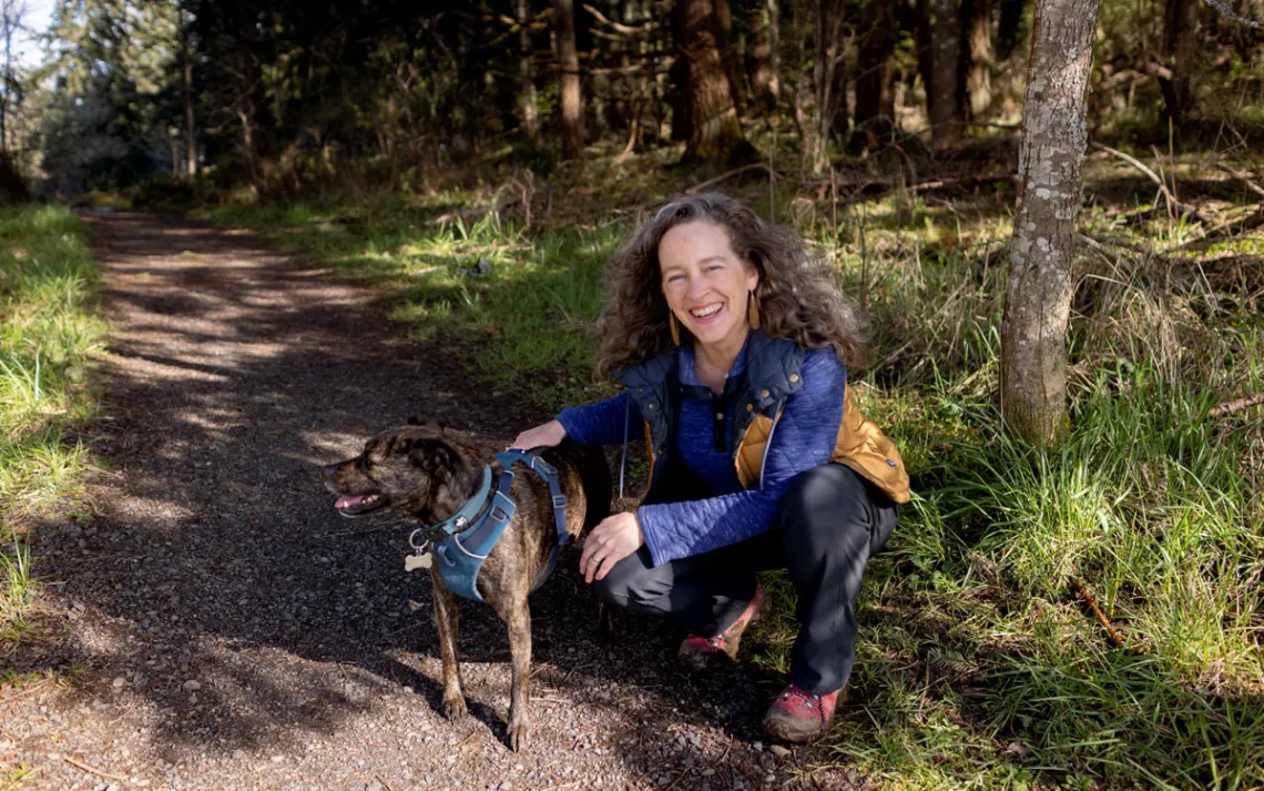  Julia Olson wears hiking clothes and smiles while kneeling down with her brown dog on a trail.