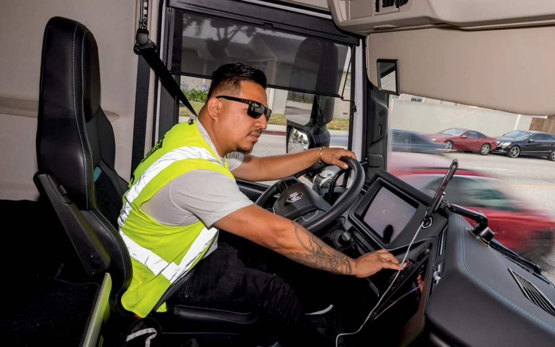Luis Cardona wears a yellow safety vest and leans forward in the driver seat of a truck
