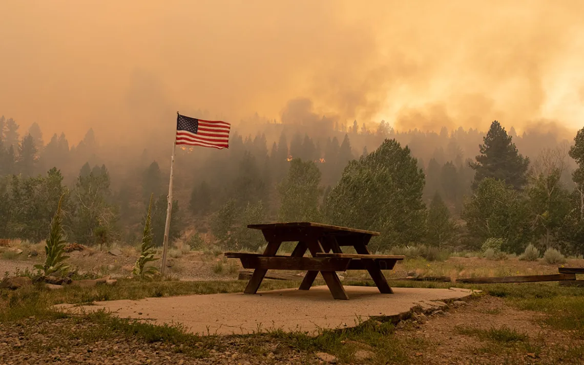 Trees burn on the horizon beyond an American flag by an empty park bench