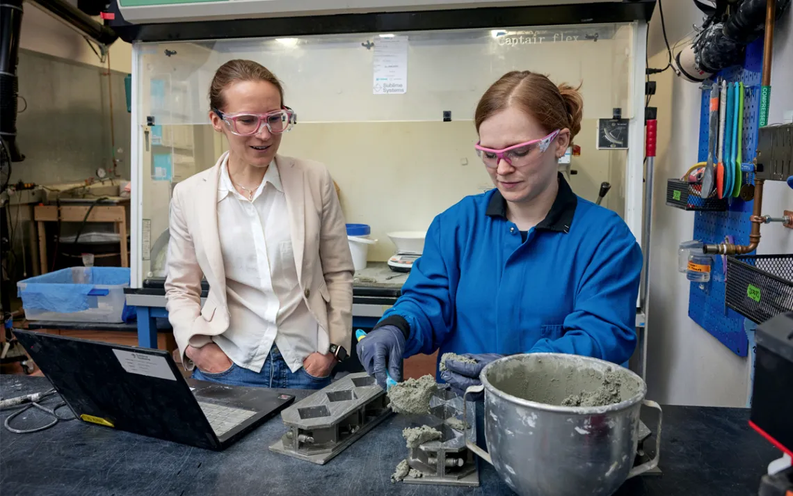 eah Ellis wears a light blazer and white button-down shirt, watching Summer Camerlo-Bass in a blue work shirt scooping gray cement from a metal mixing bowl.