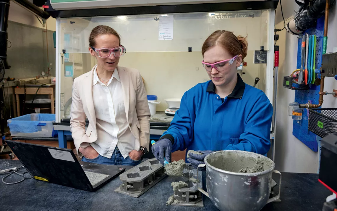 eah Ellis wears a light blazer and white button-down shirt, watching Summer Camerlo-Bass in a blue work shirt scooping gray cement from a metal mixing bowl.
